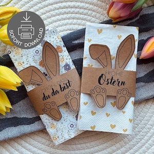 Könnte beinhalten: Zwei Servietten zum Thema Ostern mit Hasenohren- und Pfoten-Ausschnitten. Eine Serviette hat ein Blumenmuster und den Text "Schön, dass du da bist". Die andere hat goldene Herzen und den Text "Frohe Ostern".