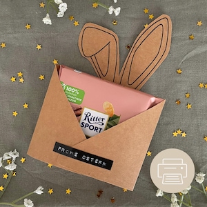 Può includere: Una busta di carta marrone con una tavoletta di cioccolato all'interno. La busta ha un ritaglio a forma di orecchio di coniglio nella parte superiore e le parole "Frohe Ostern" scritte sul davanti.