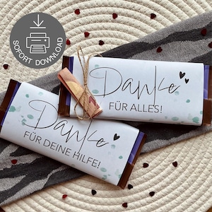 以下が含まれることがあります： 白い紙に包まれた2本のチョコレートバー。エレガントな筆記体で「Danke」と書かれています。1本のバーには、紐で結ばれた丸められた紙幣があります。「FÜR ALLES!」と「FÜR DEINE HILFE!」の文字も表示されています。ハート型の小さな紙吹雪が散らばっています。
