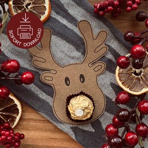Könnte beinhalten: Eine braune Rentier-Karte mit einem goldverpackten Schokoladenbonbon im Mund. Die Karte ist von roten Beeren, getrockneten Orangenscheiben und einem gestreiften grau-weißen Stoff umgeben. Auf der Karte steht "SOFORT DOWNLOAD".