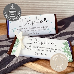 Può includere: Due barrette di cioccolato con involucri bianchi con la parola "Danke" in elegante scrittura. Una è legata con un nastro. Gli involucri includono testo in tedesco ed elementi decorativi. Le barrette sono su una superficie di legno.