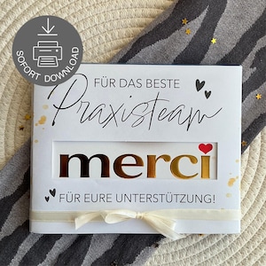 Op de afbeelding: Een witte kaart met gouden folieletters met de tekst "merci" in een uitgesneden venster. De kaart heeft de tekst "FÜR DAS BESTE Praxisteam" en "FÜR EURE UNTERSTÜTZUNG!" erop gedrukt. Onderaan een crèmekleurig lint en strik.