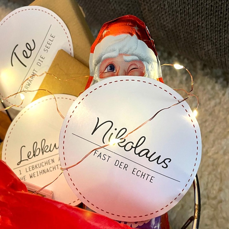 Op de afbeelding: Een kerstcadeaupakket met verschillende artikelen, waaronder een chocolade Sinterklaas, een doos thee en een Lebkuchenkoekje. De mand is versierd met een lichtsnoer en een witte cirkel met de tekst "Nikolaus Fast Der Echte".