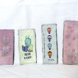 May include: Four handmade cross-stitch bookmarks. One features a basket of flowers, another a cartoon bookworm, a third with hot air balloons and the text "LET YOUR IMAGINATION SOAR 2025", and the last with a floral pattern.