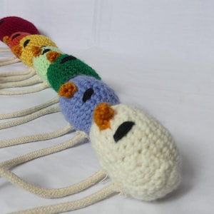May include: A colorful, handmade crochet caterpillar toy. The caterpillar is made of various colors, including red, orange, yellow, green, blue, and white. It has black eyes and orange accents. The toy is connected to a cream-colored rope.