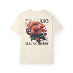 May include: Cream-colored t-shirt featuring a floral graphic of orange roses with green leaves. The design includes the text "AIC" above the flowers and "ALLINCHRIST" below, set within a black lined square.