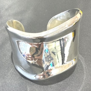 Puede incluir: Una pulsera tipo brazalete ancha de tono plateado pulido con una superficie lisa y ondulada. El metal tiene un acabado altamente reflectante que crea reflejos y sombras distorsionados en su estructura curva y abierta, formando una banda sólida.