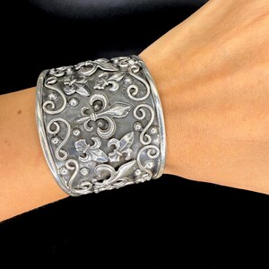 Op de afbeelding: Brede zilveren manchetarmband met een sierlijk ontwerp met lelies en krullen. De manchet is ongeveer 5 cm breed en heeft een gedetailleerd, getextureerd oppervlak. De armband wordt om de pols gedragen.