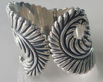 May include: A silver bracelet with a detailed, swirling, leaf-like design. The bracelet is hinged and opens to fit the wrist.