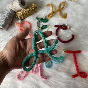 May include: A collection of colorful crocheted letter ornaments, including green, red, blue, pink, and yellow. Each letter is attached to a string with a small wooden bead.