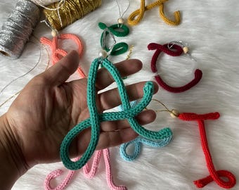 May include: A collection of colorful crocheted letter ornaments, including green, red, blue, pink, and yellow. Each letter is attached to a string with a small wooden bead.