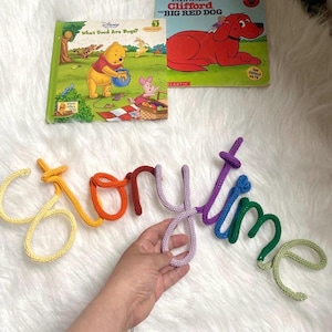 May include: A colorful, crocheted "Storytime" sign in yellow, orange, red, purple, blue, green, and light green. Two children's books are in the background, one featuring Winnie the Pooh and the other, Clifford the Big Red Dog.