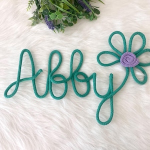 May include: A teal-colored rope name, "Abby", is displayed with a matching flower accent. The flower has teal petals and a lavender center. The name and flower are on a white, fluffy surface, with greenery in the background.