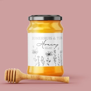 May include: A glass jar of honey with a white label that says "Zomerhuis & Tuin Honing EST. 2023" and features a black and white line drawing of flowers and bees. A wooden honey dipper is in the foreground.