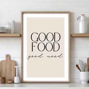 Peut inclure: Impression encadrée avec l'inscription "GOOD FOOD good mood" en lettres noires. L'impression est exposée sur un comptoir blanc avec des articles de cuisine, dont des planches à découper, une casserole et des ustensiles. Des étagères au-dessus contiennent de la vaisselle et des bocaux décoratifs.