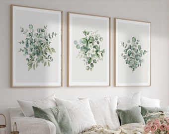 May include: Three framed watercolor prints of eucalyptus branches with white flowers, hanging on a white wall above a white couch with green throw pillows. A vase of pink flowers sits on a wooden coffee table.