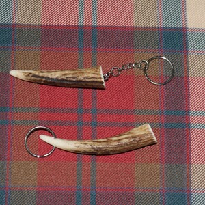 May include: Two antler keychains with silver-colored key rings. One keychain is straight, the other is curved. The antlers are brown and cream-colored. The keychains are on a red, blue, and tan plaid background.