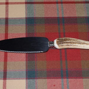 May include: A cake server with a serrated edge and a dark blade. The handle is made of a light-colored antler material. The server is set against a red, beige, and green plaid background.