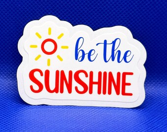 May include: A white die-cut sticker featuring a yellow sun icon with a red center, the blue cursive text "be the" and the red uppercase text "SUNSHINE". The sticker has a white border and is presented against a solid, vibrant blue background.