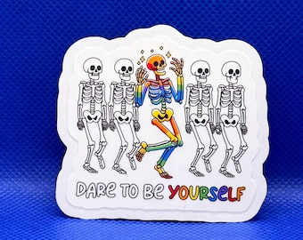 May include: A white die-cut sticker featuring four black and white skeletons standing in a row with a central rainbow-colored skeleton dancing. The text below reads DARE TO BE YOURSELF with the word YOURSELF in rainbow colors.