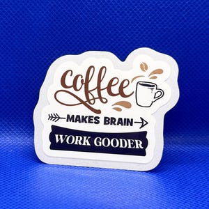 May include: A white sticker with the text "coffee makes brain work gooder" in a mix of fonts and colors. The word "coffee" is in a brown script font, and the phrase is accompanied by a coffee cup illustration.