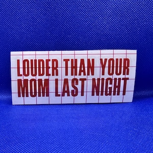 May include: A white rectangular sticker with red text that reads "LOUDER THAN YOUR MOM LAST NIGHT." The sticker has a grid pattern and is set against a blue background.