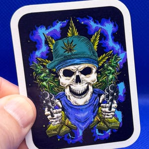 May include: A sticker featuring a skull wearing a teal cap with a cannabis leaf, a blue bandana, and holding two revolvers. The background includes cannabis leaves and blue smoke against a black backdrop.