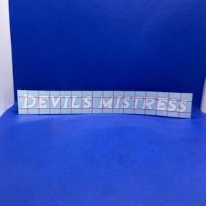 May include: A rectangular sticker with the text "DEVIL'S MISTRESS" in white, block letters. The sticker has a light blue background with a grid pattern. The sticker is placed on a blue surface.