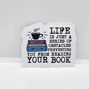 May include: White sticker with a quote about reading. The design includes a stack of books, a steaming coffee cup, and the text "LIFE IS JUST A SERIES OF OBSTACLES PREVENTING YOU FROM READING YOUR BOOK."