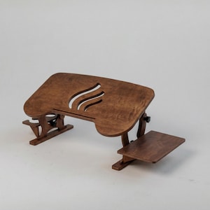 Puede incluir: Un escritorio para portátil de madera marrón con una plataforma principal ajustable que presenta tres aberturas de ventilación curvas y una bandeja lateral más pequeña. El escritorio se apoya en dos patas plegables con perillas negras.