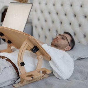 Może przedstawiać: Podstawka pod laptopa z jasnego drewna z regulowaną platformą, czarnymi metalowymi zawiasami i dwoma okrągłymi wycięciami w bocznej podstawie. Podstawka została zaprojektowana tak, aby bezpiecznie utrzymywać laptopa nad płaską powierzchnią.