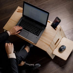 Puede incluir: Una persona trabajando en un ordenador portátil sobre un escritorio de madera. El escritorio tiene un soporte para teléfono incorporado y una alfombrilla para el ratón. El portátil está abierto y la persona está escribiendo en el teclado. El teléfono está en el soporte y el ratón está en la alfombrilla.