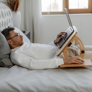 Puede incluir: Un soporte ajustable para computadora portátil hecho de madera clara con un marco de soporte curvo y una base plana para el mouse. El soporte cuenta con un mecanismo de bisagra con perillas negras para ajustar el ángulo, sosteniendo una laptop plateada.