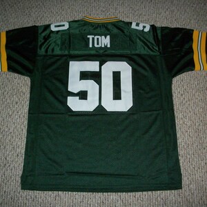 May include: Green football jersey with white lettering. The number 50 is on the back of the jersey with the name TOM above it.