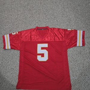 May include: A red football jersey with the number 5 in white. The jersey has white and yellow stripes on the sleeves. The jersey is made of a shiny material.