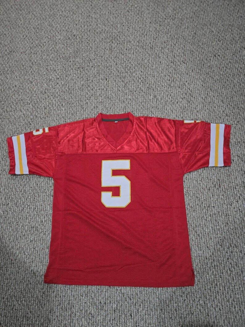 May include: A red football jersey with the number 5 in white. The jersey has white and yellow stripes on the sleeves. The jersey is made of a shiny material.