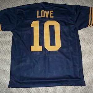 Puede incluir: Camiseta de fútbol azul marino con letras y números dorados. La palabra "LOVE" está encima del número "10" en la espalda. Rayas doradas adornan las mangas. La camiseta está hecha de tejido de malla transpirable.