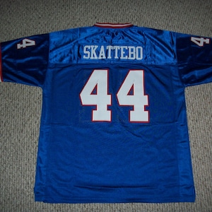 Pode incluir: Uma camisola azul com o nome "SKATTEBO" nas costas superiores e o número "44" em branco com acabamento vermelho. As mangas têm o número "4" e riscas vermelhas e brancas.