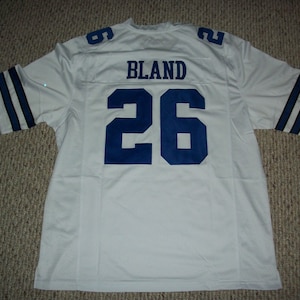 May include: White football jersey with blue and white striped sleeves. The back of the jersey has the name "BLAND" and the number "26" in blue.