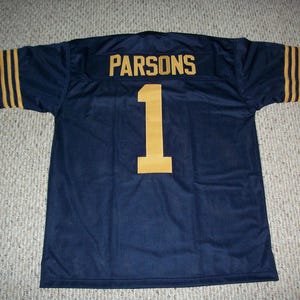 Puede incluir: Camiseta de fútbol azul marino con letras y números dorados. El nombre "PARSONS" está en la parte superior de la espalda, con el número "1" en dorado debajo. Rayas doradas acentúan las mangas. La camiseta es de tejido de malla transpirable.