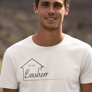 Könnte beinhalten: Weißes T-Shirt mit einer schwarzen und grauen Hausgrafik und dem Text "est. 2024 Bauherr" auf der Vorderseite.