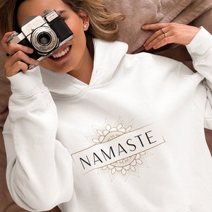 Peut inclure: Sweat-shirt blanc avec un motif de mandala doré et le mot "NAMASTE" en texte noir.