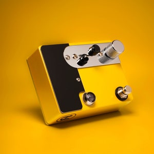 Può includere: Un pedale per effetti per chitarra giallo e nero con due manopole e un interruttore a pedale. Il pedale ha una piastra cromata con una manopola nera e una manopola argentata.