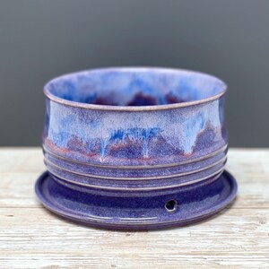 May include: A cylindrical ceramic planter with a glossy glaze featuring a blend of deep purple, violet, and light blue tones. The exterior has three horizontal ridges around the middle, and the attached saucer base includes a small drainage hole.