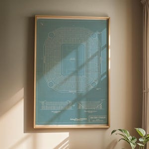 May include: A framed blueprint of a stadium, featuring detailed architectural drawings in white against a teal background. The frame is a light wood color. The artwork hangs above a sofa with green, tan, and teal pillows.