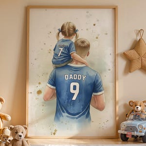 May include: A framed watercolor print depicts a father with a child on his shoulders, both wearing blue jerseys with the word "DADDY" and the number 9. The artwork is surrounded by teddy bears, star-shaped decorations, and a toy car.