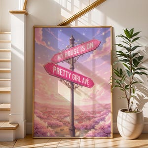 Puede incluir: Una impresión artística enmarcada que presenta un letrero de calle caprichoso con las frases "MY HOUSE IS ON" y "PRETTY GIRL AVE" en flechas rosas. La obra de arte representa un paisaje de ensueño con un cielo rosa y púrpura, y está enmarcada en madera clara.