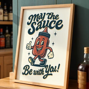 Op de afbeelding: Een ingelijste print met de tekst "May the Sauce Be with You!" toont een cartoon ketchupfles karakter die een duim omhoog geeft. De print heeft een licht houten frame en staat tegen een neutrale achtergrond.