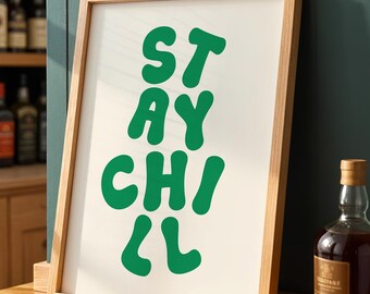 May include: A framed art print with the words "STAY CHILL" in bold, green letters. The print has a cream-colored background and a natural wood frame. The print is displayed on a wooden surface, with bottles of liquor in the background.