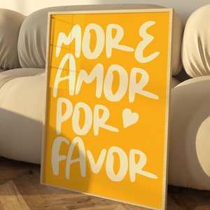 May include: A yellow framed poster with white text that reads "More Amor Por Favor" with a small heart symbol. The poster is leaning against a white sofa.